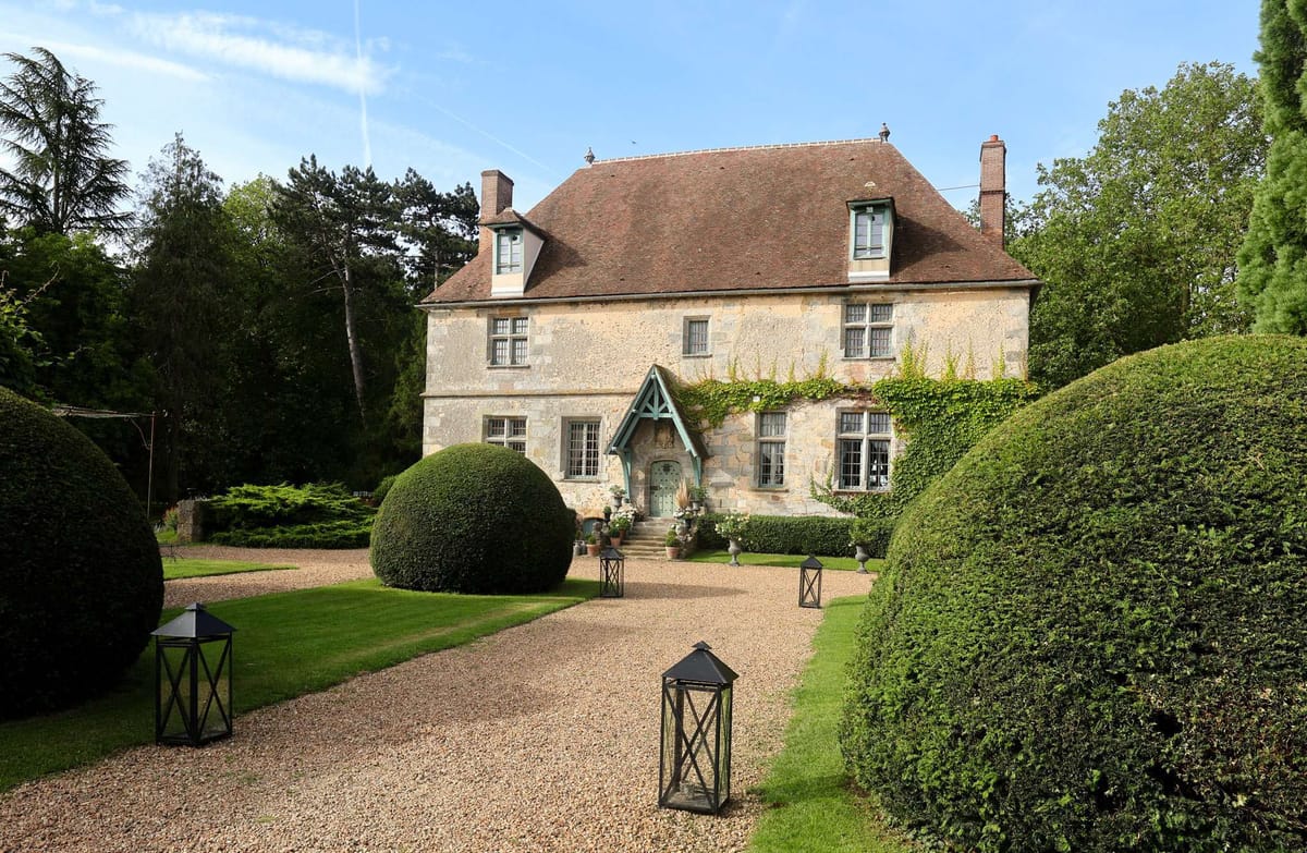 A wide exterior shot of a French country manor house featuring a honey-colored stone façade, terracotta-tiled mansard roof with dormer windows, and a distinctive entrance porch with pale blue-green timber framing covered in climbing vines. The formal front garden includes neatly clipped spherical and rounded box topiary flanking a gravel driveway, with black metal lanterns placed along the path leading to the entrance. White floral arrangements in pots are visible on the front steps, adding a decorative wedding touch. No people are visible in the frame. Potential venue feature image.