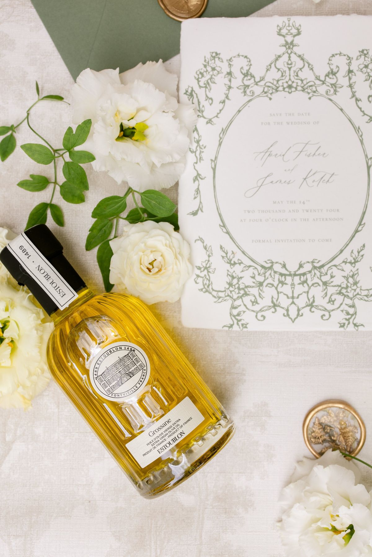 Flat lay of save-the-date card with rococo frame, olive oil bottle, white roses and peonies on cream linen