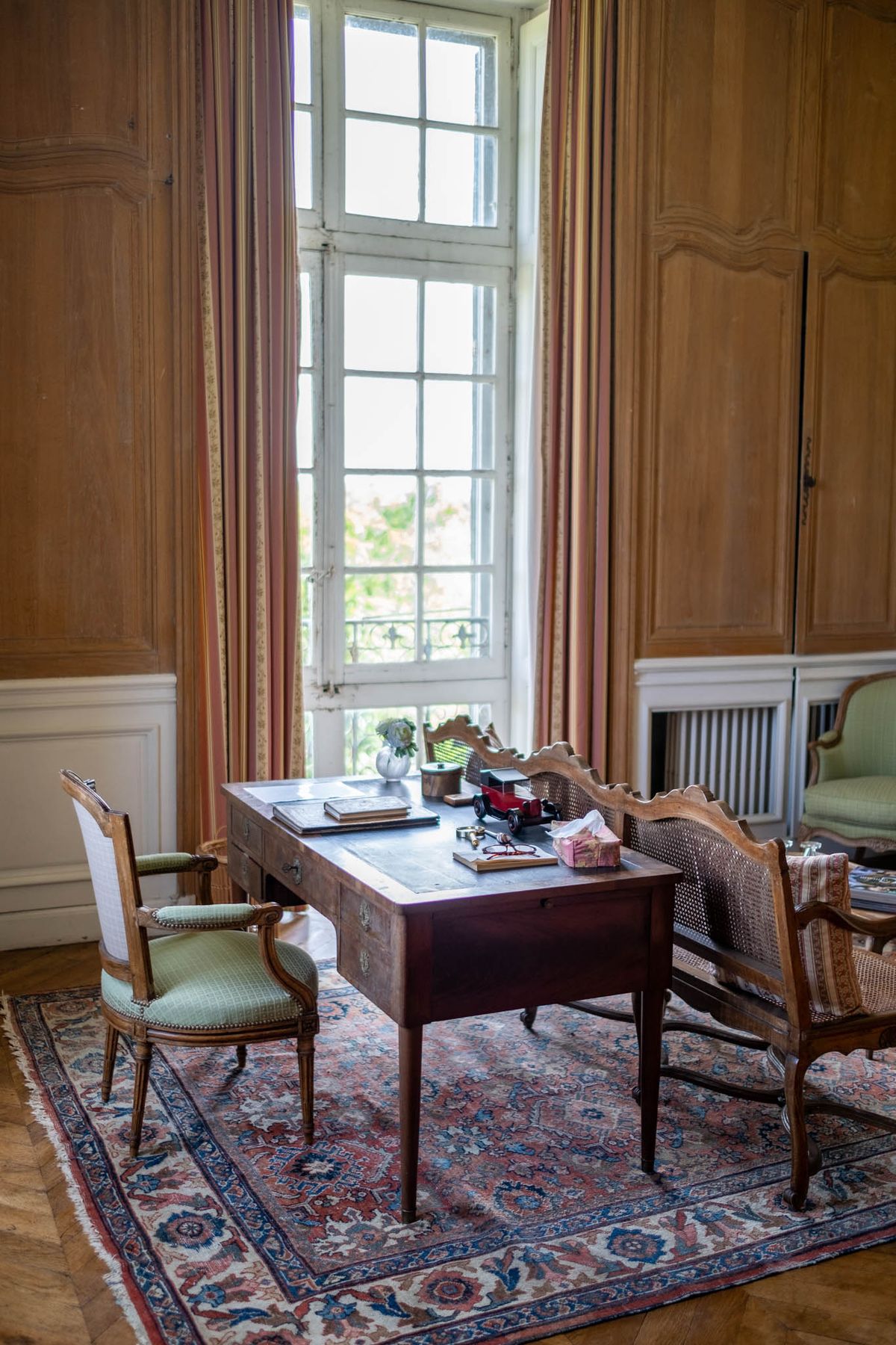 Interior room shot of a château study or salon, with no people present. The room features floor-to-ceiling warm honey-toned wood panelling, a tall multi-pane white-framed French window with salmon and gold striped curtains, and a herringbone parquet floor. A dark mahogany writing desk sits at the centre on a large Persian-style rug in dusty rose, navy blue, and ivory tones. Around the desk are two Louis XVI-style armchairs with sage green upholstered seats and a cane-backed settee. The desk surface holds personal items including books, a small red toy car, a small white vase with greenery, and a gift box. A cast-iron radiator is visible in the background alongside a partially visible green sofa. The composition is a wide interior shot taken from a slightly elevated angle. Potential venue feature image.