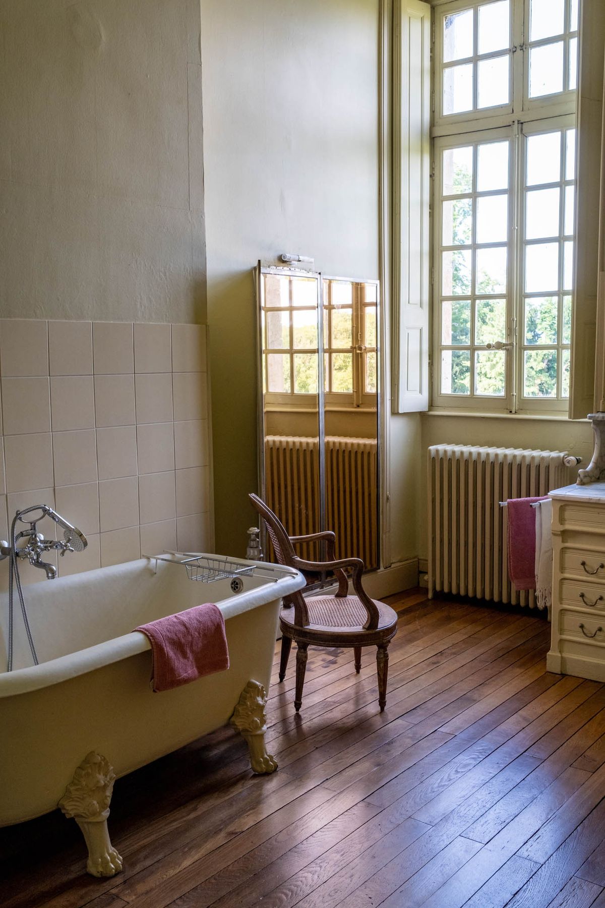 Chateau bathroom with cream clawfoot tub Louis XVI cane chair tall French windows and dark hardwood floors