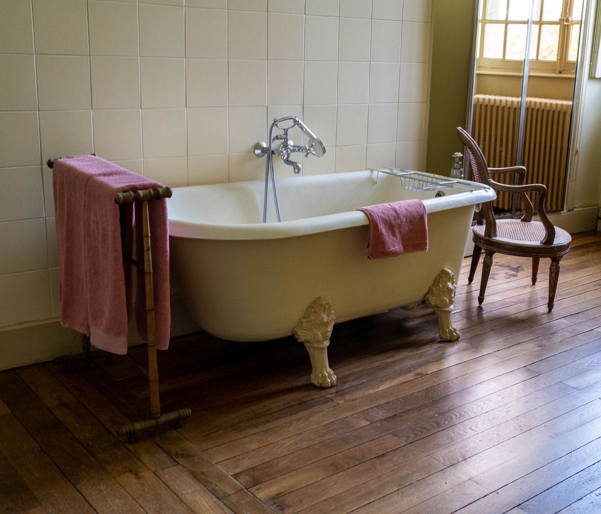 This image shows a bathroom interior featuring a cream-painted cast-iron clawfoot bathtub with ornate lion's paw feet and chrome wall-mounted faucet with handheld shower attachment. A metal bath caddy rests across the tub's rim, and dusty rose-pink towels are draped over both the tub's edge and a freestanding wooden towel rail to the left. A vintage cane-seat French-style armchair sits to the right of the tub, and a cast-iron radiator is visible near a multi-pane window in the background. The room has wide-plank dark wood flooring, cream square wall tiles on the lower half, and an olive-green painted upper wall, giving the space a classic French country house character. Potential venue feature image.