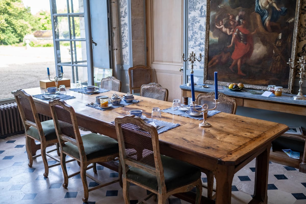 An interior dining room at what appears to be a French château, shown without any wedding party or guests present. A large rustic oak farmhouse table is set for approximately six people using blue-and-white porcelain teacups and saucers, woven navy blue placemats, silver candle holders with tall cobalt blue taper candles, and glass tumblers. The chairs are Louis XV-style carved oak with cane backs and green velvet seat cushions. The room features a black-and-white toile de Jouy wallpaper panel, a large oil painting in a gilt frame depicting a classical figurative scene, a blue-grey marble console table along the wall, and a geometric blue-and-white diamond-patterned tile floor. Tall French doors open onto a courtyard. The overall decor palette is navy blue, white, and warm wood tones in a classic French château interior setting. Wide-angle interior shot. Potential venue feature image.