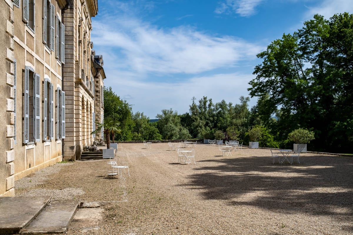 An outdoor terrace or gravel courtyard alongside a large French château, set up for a cocktail hour or reception gathering. The château facade features pale stone construction with grey-blue shuttered windows across multiple floors. White wrought-iron bistro tables and chairs are arranged informally across the gravel terrace, with large white square planters positioned along the perimeter. No people are present in the image; this appears to be a venue setup shot taken in daytime. The composition is a wide horizontal shot taken from ground level, showing the full length of the terrace receding into the distance. Potential venue feature image.