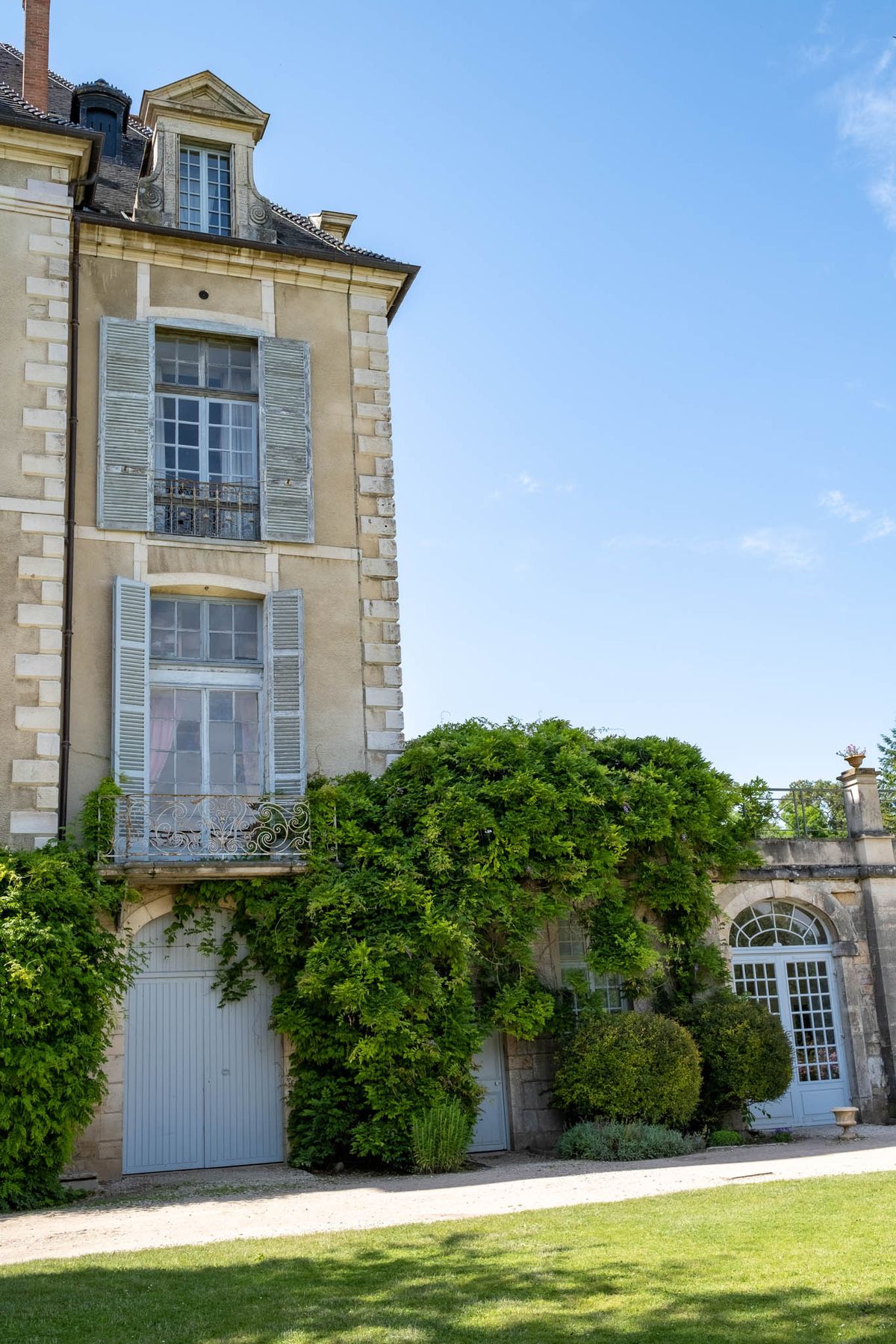 An exterior architectural shot of a French château, showing a multi-storey stone façade with pale blue-grey wooden shutters, ornate wrought-iron balconies, and a mansard roof with a dormer window. The ground floor features pale blue painted doors partially covered by climbing greenery, and an adjoining lower wing with an arched Palladian-style doorway in white. The composition is a wide shot taken from ground level at a slight angle, capturing the building's side elevation alongside a gravel path and trimmed lawn. No people are present in the image. Potential venue feature image.