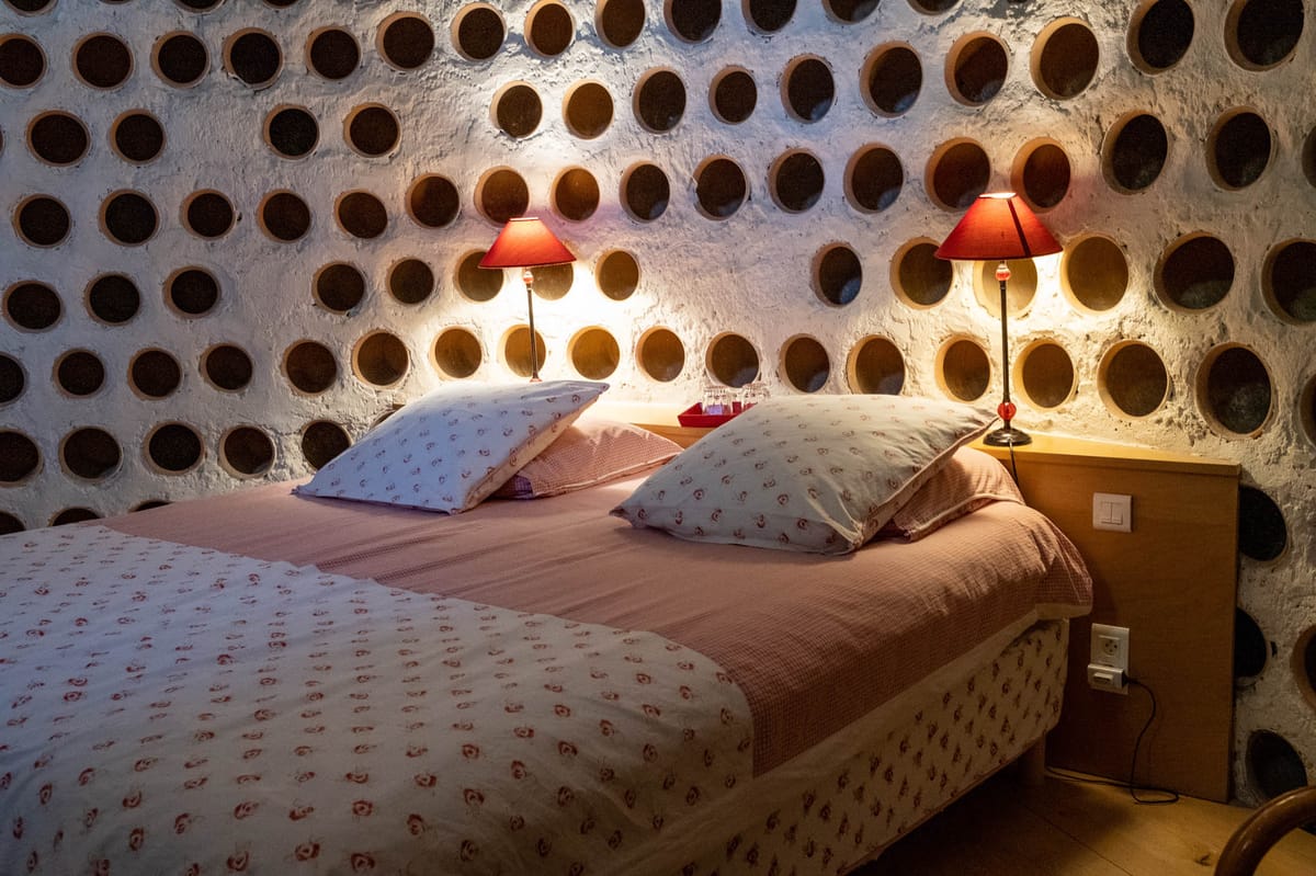 Wine-cave bedroom with circular bottle niches, white plaster walls, rose bedding, and red-shaded lamps