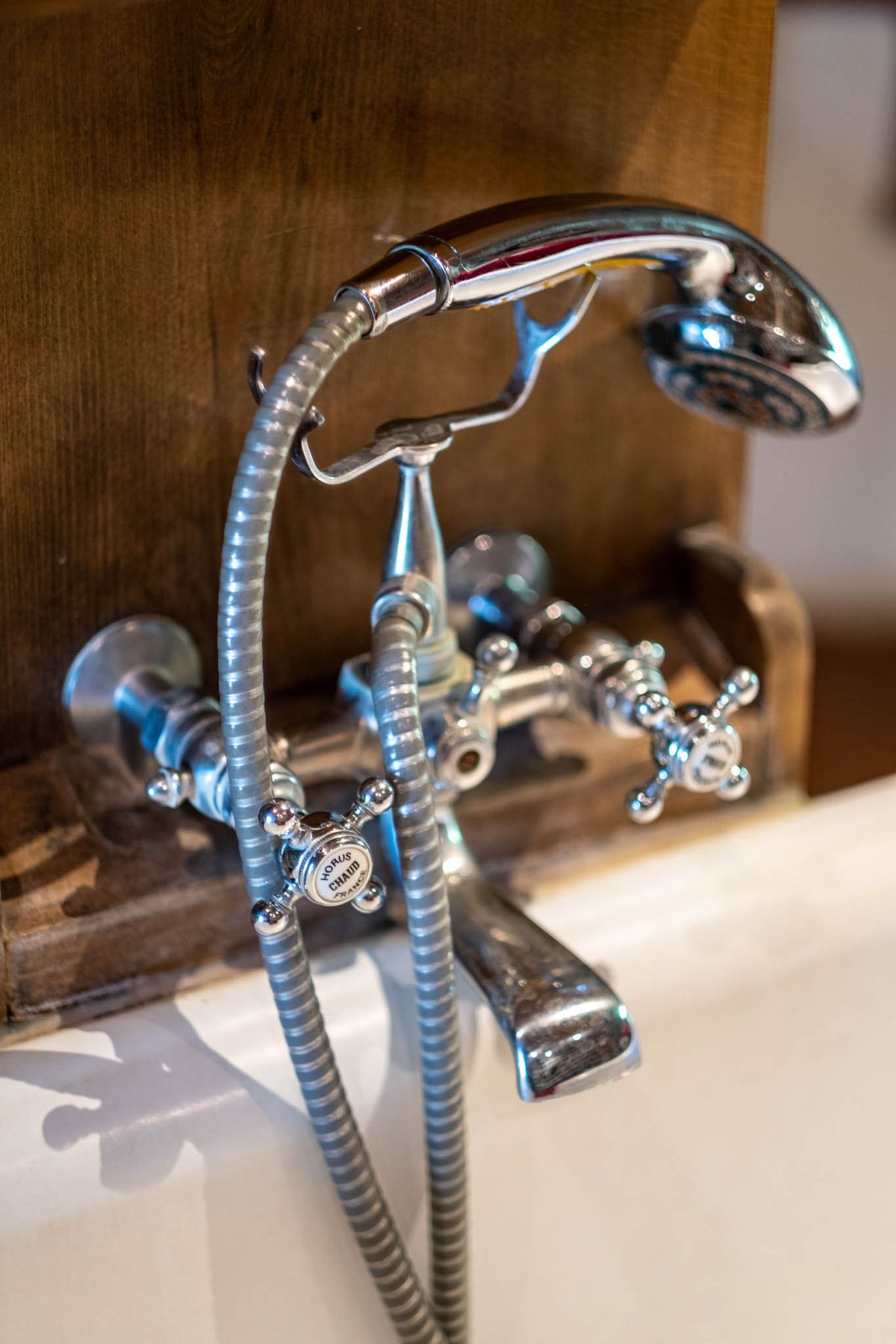 Close-up detail shot of a vintage-style chrome bath faucet set mounted on the edge of a white bathtub, with cross-handle taps labeled 'Chaud' (hot) in French, a handheld shower head, and a flexible chrome hose. The fixture has a classic, period-inspired design with polished chrome finish. The background features rustic raw wood paneling, contrasting with the bright metalwork. This image appears to be a venue accommodation or bathroom detail shot rather than a wedding photograph.
