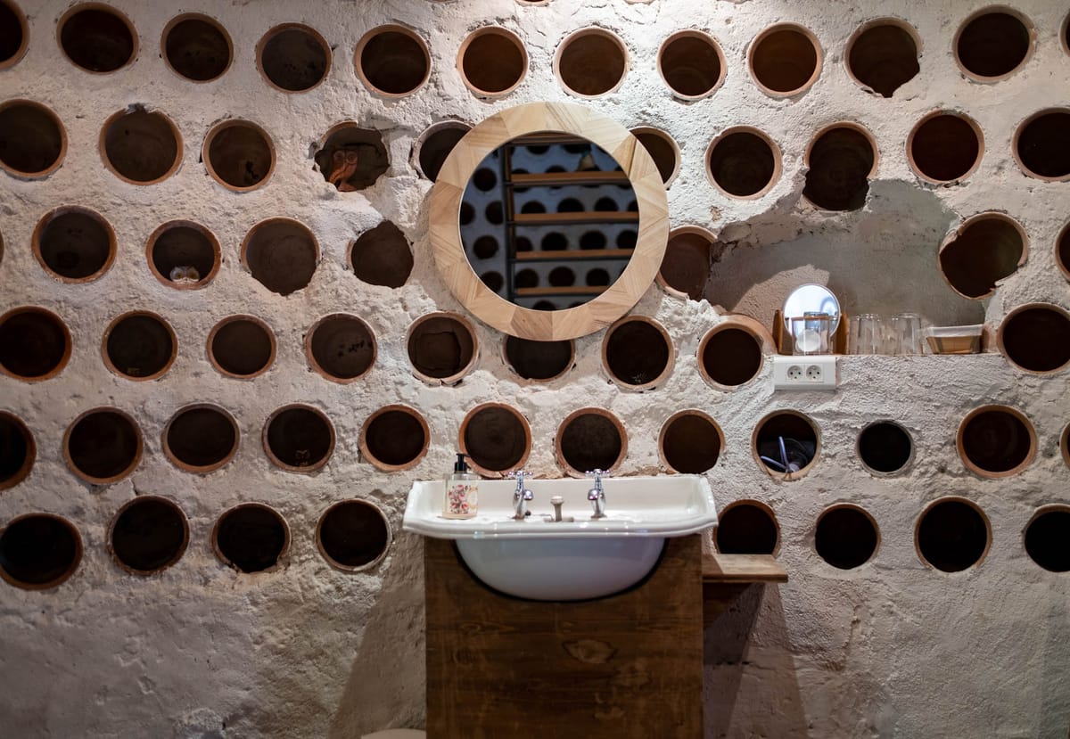 Venue bathroom with terracotta wine bottle cavity wall, white basin on raw wood vanity, and geometric wood-framed mirror