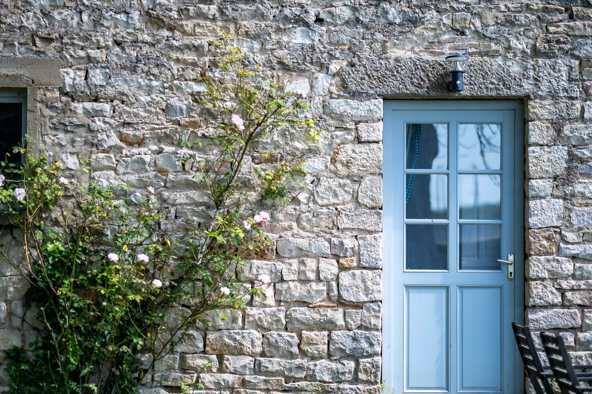 A close-up architectural shot of a rustic French country building exterior, showing a pale blue-grey painted door with six glass panes in the upper half and two raised panels below, fitted with a silver lever handle and a black wall-mounted lantern above. The facade is built from irregular cut limestone blocks in warm grey and cream tones. A climbing rose bush with small blush pink blooms grows against the left side of the wall. The edge of a dark metal chair is visible at the lower right corner. No people are present in the frame. Potential venue feature image.