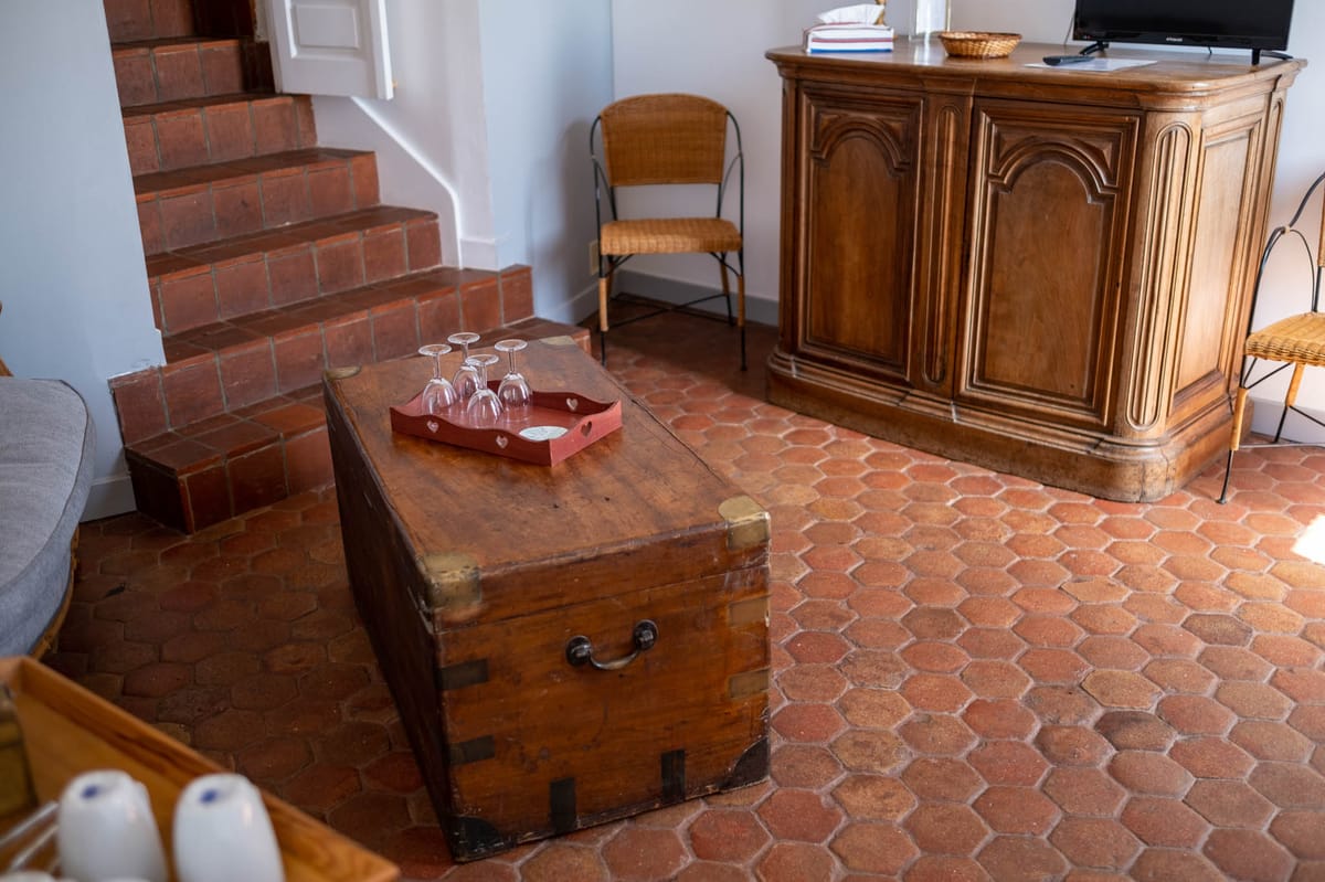 Rustic French country living room with terracotta floor tiles, wooden trunk, carved armoire, and brick staircase