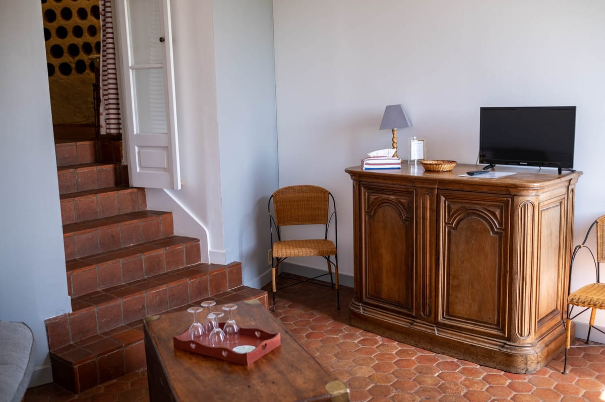 An interior room shot of what appears to be a guest accommodation or gîte on a French wedding venue property. The room features terracotta hexagonal floor tiles, a carved wooden armoire topped with a small grey lamp, a wicker basket, folded linens, and a small flat-screen television. A rustic wooden coffee table in the foreground holds a red painted tray with several wine glasses. Terracotta brick steps lead up to an adjoining room through a white-painted doorway, with a glimpse of a red and white striped curtain visible beyond. A wicker and metal chair sits against the pale grey wall. The overall style is classic French provincial with a mix of antique and practical furnishings. Wide interior shot. Potential venue feature image.