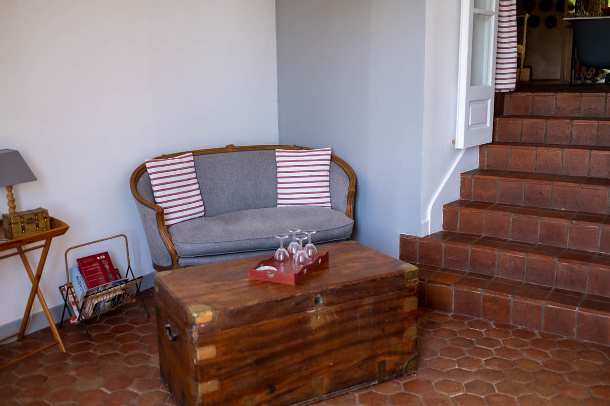 Interior seating area of a French country-style venue, showing a grey upholstered Louis XV-style wooden settee with red and white striped cushions placed against a pale blue-grey wall. A vintage wooden trunk with brass hardware serves as a coffee table, topped with a red tray holding several empty wine glasses. The terracotta hexagonal tile floor and brick staircase on the right add to the rustic French country aesthetic, while a side table with a small lamp and a magazine rack are visible to the left. Wide shot with no people present. Potential venue feature image.