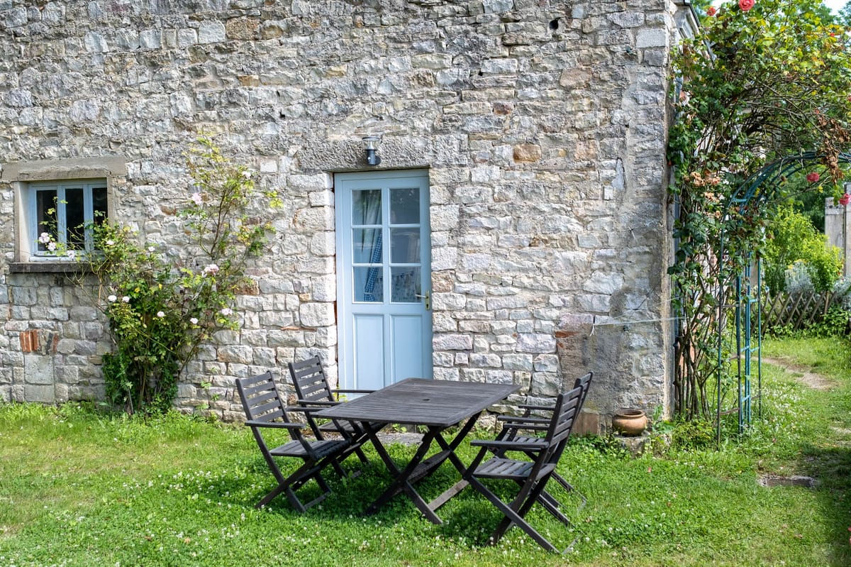 An outdoor seating area in front of a French rural stone building, featuring a dark wood folding table with four matching folding chairs set on a lawn. The building facade is constructed from irregularly cut limestone blocks with a pale blue-painted door and a small window with a matching blue frame. A climbing rose bush with pale pink blooms grows against the wall to the left of the door, and a teal metal garden arch with additional roses is visible to the right. No people, wedding party, or wedding-specific decor are present in this image. Wide shot, natural daylight. Potential venue feature image.