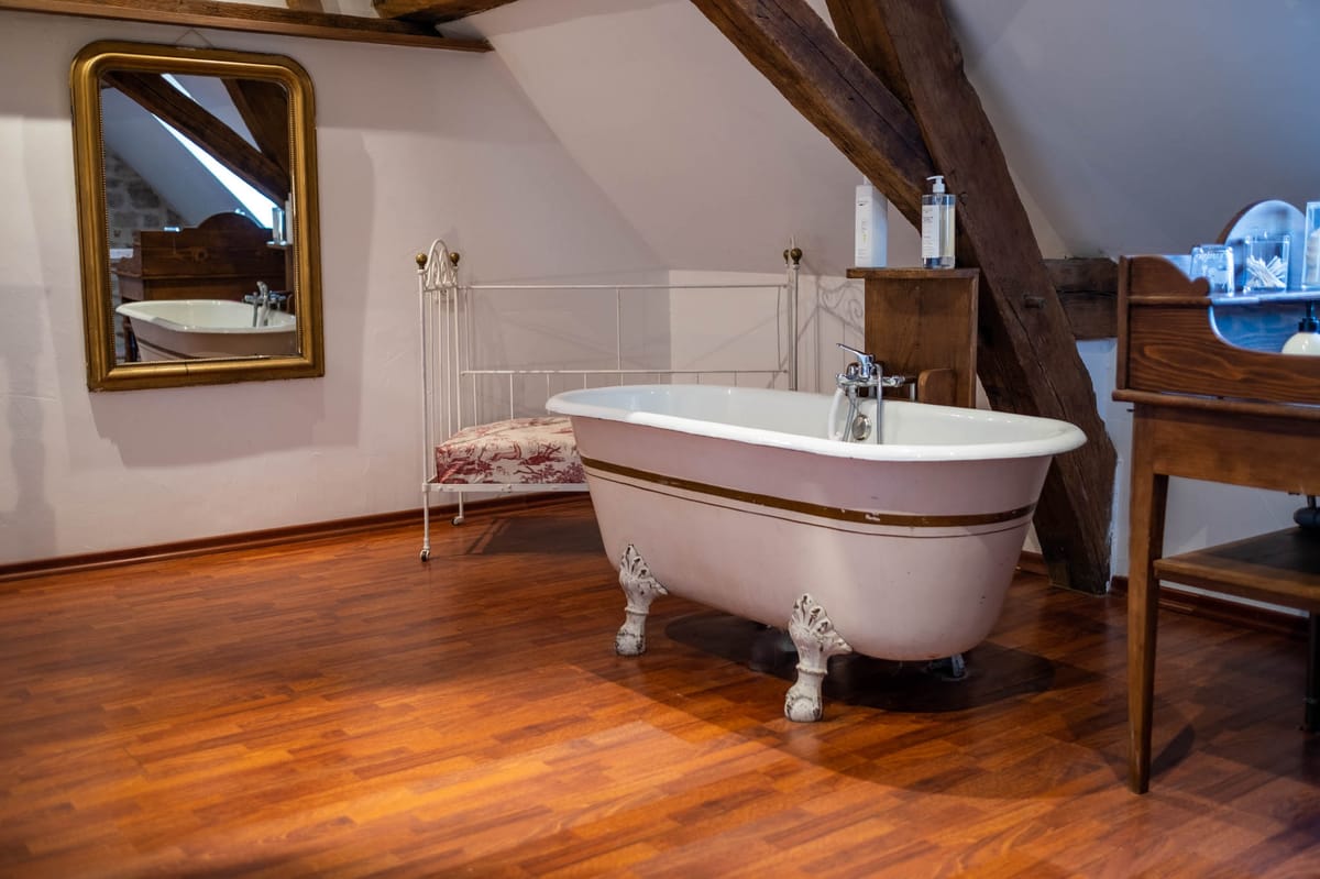 Interior shot of a getting-ready suite or bridal room, likely located in a French château or manor house. The room features a freestanding white clawfoot bathtub with a gold trim stripe and chrome faucet, positioned on warm-toned hardwood floors beneath exposed wooden roof beams in an attic-style space. A gold-framed wall mirror, a white wrought-iron chair with a red and white toile de Jouy cushion, and a dark wood washstand with toiletry products complete the styling, which mixes rustic and classic French country aesthetics. Wide-angle interior shot with no people present. Potential venue feature image.