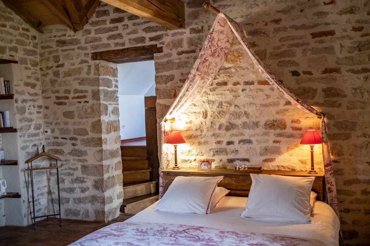 Rustic bedroom with exposed stone walls, terracotta tiles, and red toile de Jouy draped canopy bed