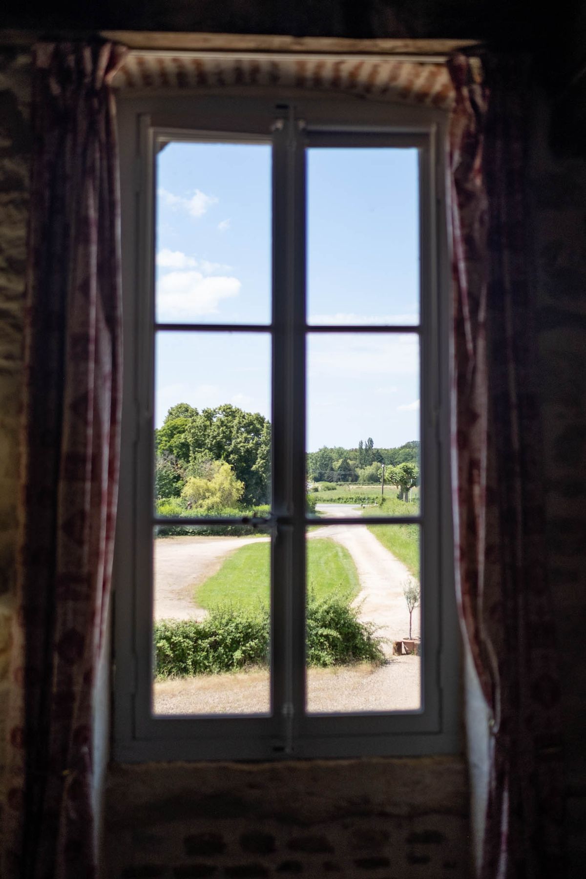 View through tall white casement window with toile curtains to gravel driveway and countryside beyond