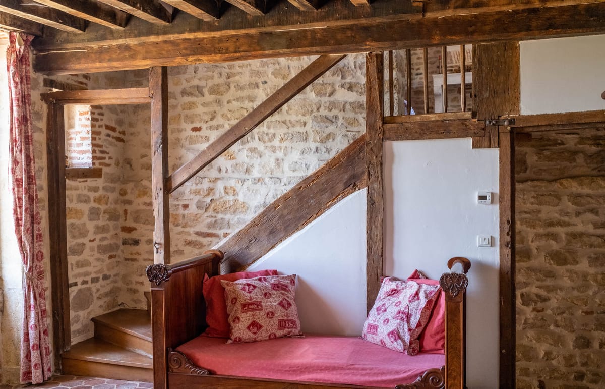 Rustic timber-frame room with stone walls carved daybed raspberry cushion and toile de Jouy curtain