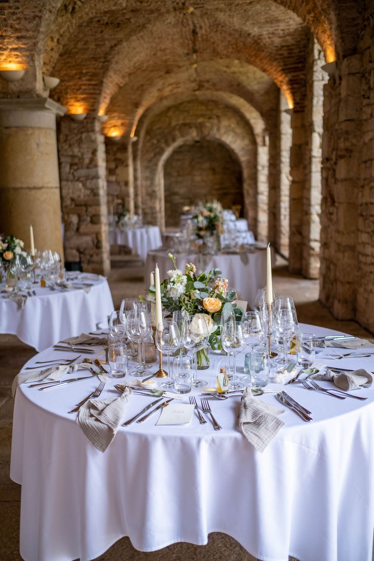A wedding reception setup photographed indoors within a historic vaulted stone hall featuring exposed brick arched ceilings and warm amber uplighting along the columns. The foreground shows a round guest table dressed in a white floor-length linen tablecloth, set with multiple crystal wine glasses, silver cutlery, and textured linen-check napkins folded at each place setting; a calligraphy place card is visible at the nearest setting. The low centerpiece features peach garden roses, ivory roses, white lisianthus, and eucalyptus foliage, flanked by two tall ivory taper candles on brass candlestick holders. Several additional round tables with matching white linens, floral centerpieces, and candlesticks are visible in the background, receding toward a head table against the far wall. The overall decor palette is white, ivory, peach, and soft gold against the warm-toned historic architecture. Wide interior shot with slight depth of field. Potential venue feature image.