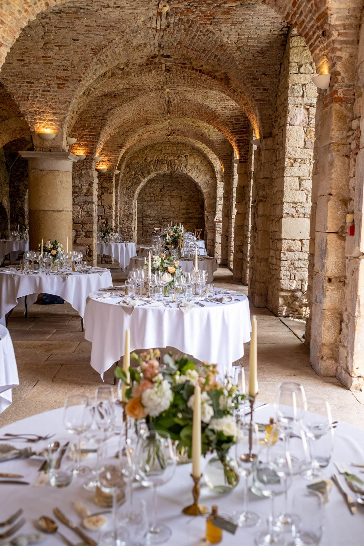 Reception tables in vaulted stone cellar with brick arches, cream hydrangea centerpieces, gold candlesticks, and crystal g...