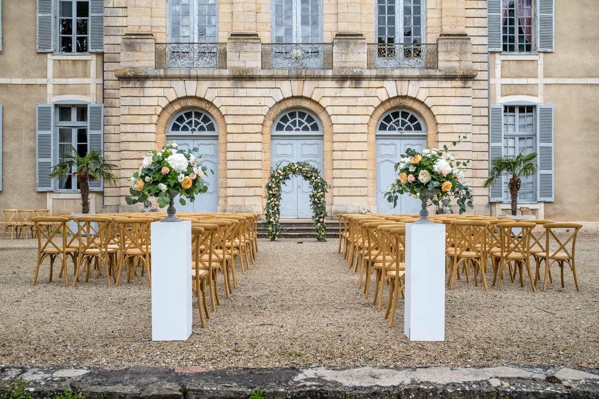 Outdoor ceremony with cross-back chairs, hydrangea urns, and floral arch at chateau entrance