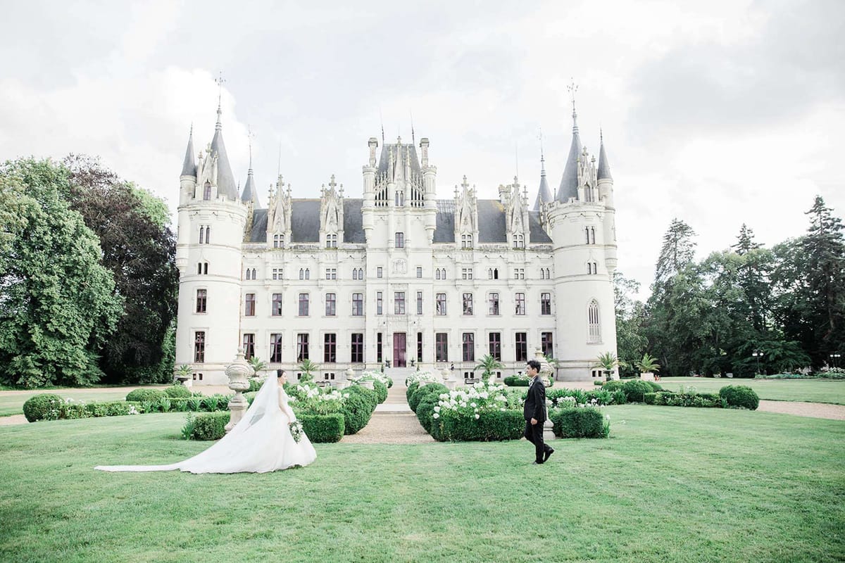 A bride and groom are photographed walking toward each other on the front lawn of a large French château, captured in a wide shot. The bride wears a white ballgown with a very long cathedral-length veil trailing across the lawn, and carries a white and green bouquet, while the groom is dressed in a black tuxedo. The château features white stone Gothic Revival architecture with pointed turrets, ornate façade detailing, and a dark slate roof, flanked by formally clipped boxwood hedging and white flowering plants along the central pathway. The composition places the couple small against the scale of the building, emphasizing the grandeur of the architecture and the formal garden layout. Potential venue feature image.