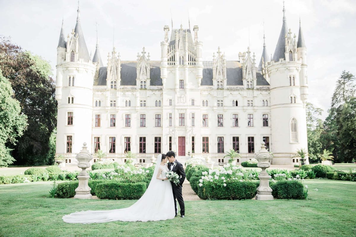 A couple portrait taken outdoors on the manicured lawn directly in front of a large white French Gothic château featuring multiple pointed turrets, dark slate roofing, ornate stone detailing, and deep red-trimmed windows and doors. The bride wears a white ball gown with an off-shoulder neckline and an exceptionally long cathedral-length veil that extends across the lawn, and she holds a bouquet of white peonies and greenery. The groom wears a black tuxedo with a black bow tie. The two stand forehead-to-forehead in a close romantic pose. The formal garden between them and the château includes neatly trimmed boxwood hedges, stone urn pedestals, and white flowering plants. Wide shot, capturing the full façade of the château and the full length of the bride's train. Potential venue feature image.