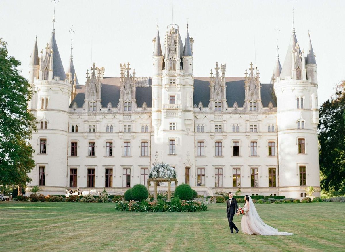 Bride in blush ballgown and groom in tuxedo posing on lawn before a white Gothic-Renaissance chateau with turrets