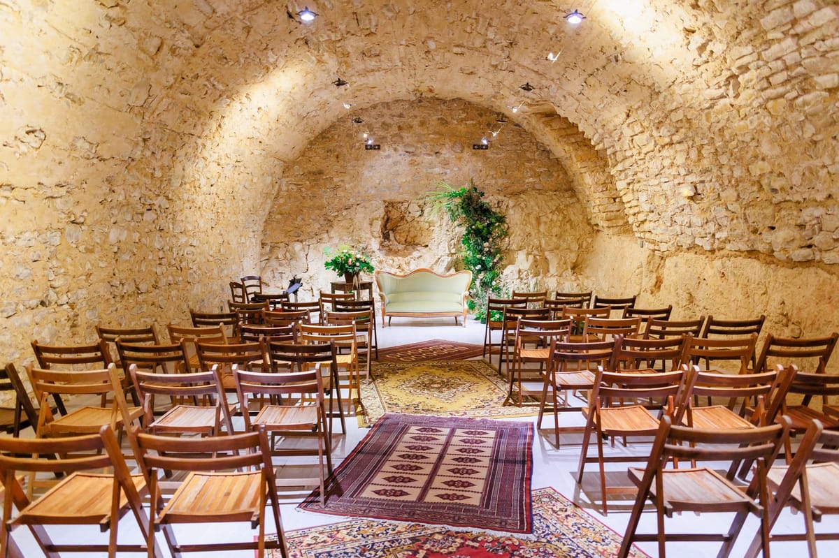 An indoor ceremony space set up inside a vaulted stone cave or underground cellar, photographed as a wide shot with no guests or couple present. Rows of wooden folding chairs are arranged on either side of a central aisle, which is lined with layered vintage-style rugs in deep red, burgundy, and golden yellow tones. At the far end of the aisle, a pale mint green upholstered vintage sofa serves as the focal point, flanked by tall greenery arrangements featuring lush tropical leaves and small floral accents in warm orange and cream tones. Spotlights mounted to the vaulted ceiling illuminate the warm-toned limestone walls. The overall styling is bohemian with vintage touches, combining the ornate sofa, layered rugs, and abundant greenery against the raw architectural backdrop. Potential venue feature image.