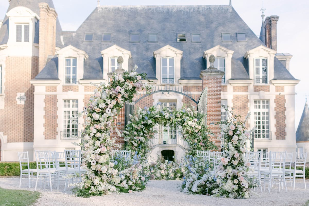 An outdoor wedding ceremony setup photographed in a wide shot in front of a French château with red brick and stone façade, mansard roof, and ornate ironwork gate. The ceremony features two large circular floral arches positioned at the altar, densely covered in white garden roses, blush pink roses, white anemones with dark centers, soft lavender blooms, blue delphinium, and lush green foliage. Additional floral ground arrangements cascade at the base of each arch and along the aisle, continuing the same soft pastel palette of blush, white, lavender, and powder blue. White chiavari chairs are arranged in rows on either side of a gravel aisle, set up to accommodate approximately 40–60 guests. The overall styling is classic and romantic with a soft pastel floral palette, set entirely outdoors on a gravel courtyard directly in front of the château entrance. Potential venue feature image.