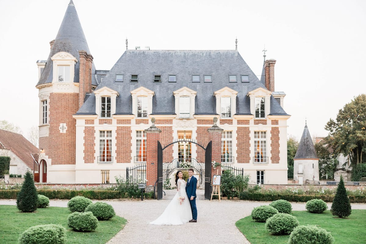 A couple poses for a portrait in front of a French château, standing on a gravel path that leads to ornate black iron gates at the château entrance. The bride wears a long-sleeved lace ball gown with a cathedral-length train and carries a soft pink and white bouquet, while the groom wears a navy blue suit. The château is a red brick and cream stone building with a slate mansard roof, cone-topped turrets, and tall white-trimmed windows; warm interior lighting is visible through the ground-floor windows. A wooden easel with a welcome sign stands to the right of the couple near the gates, which are decorated with a white floral arrangement. The formal garden in the foreground features clipped spherical and conical topiary shrubs arranged symmetrically along the path. This is a wide shot taken at dusk or in soft overcast light. Potential venue feature image.