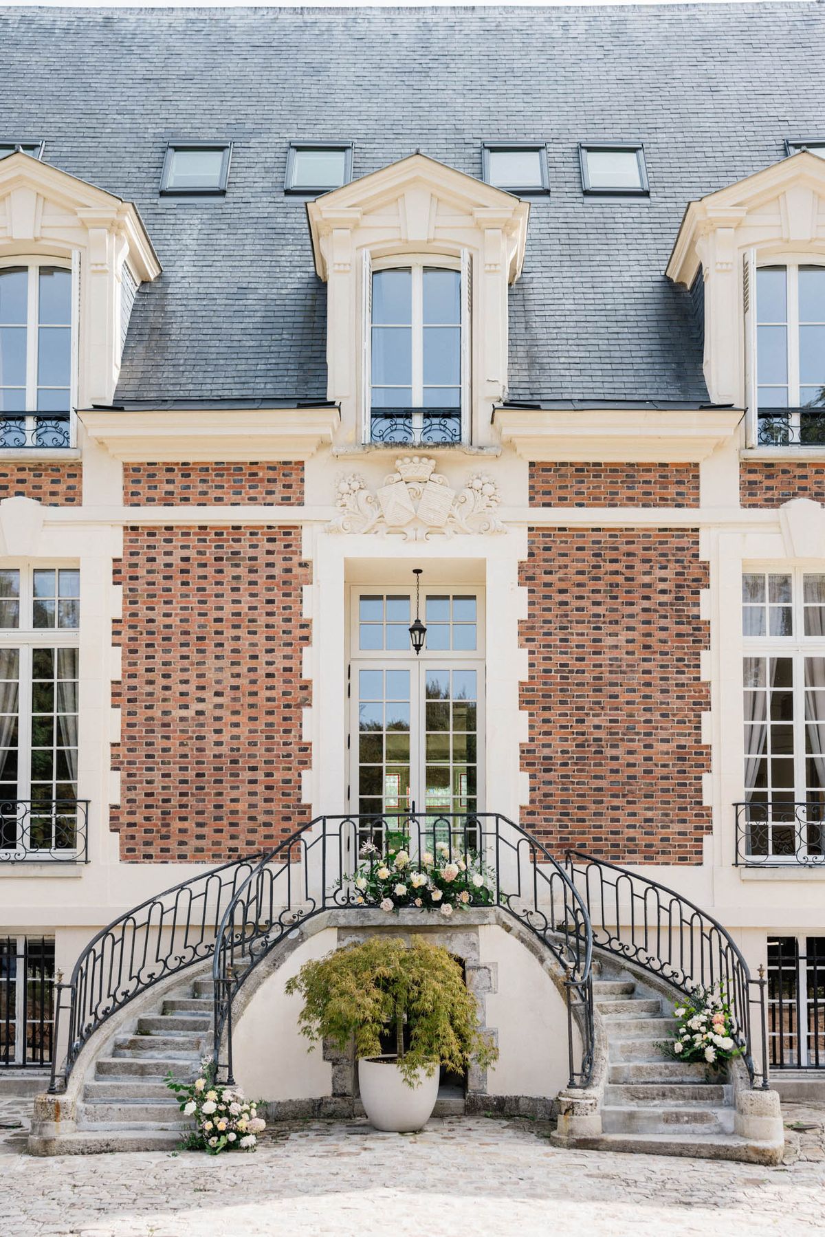 A wide exterior shot of a French château facade featuring red brick with cream stone quoins, a slate mansard roof with dormer windows, and ornate wrought-iron balconies. The entrance is framed by a symmetrical double horseshoe staircase with curved wrought-iron railings, decorated with blush and ivory floral arrangements — including roses and mixed blooms — placed at the base of each staircase and at the landing. A large potted ornamental shrub sits beneath the central arch of the staircase, and a hanging lantern is visible above the entrance door. The cobblestone forecourt is visible in the foreground. No people are present in the image. Potential venue feature image.
