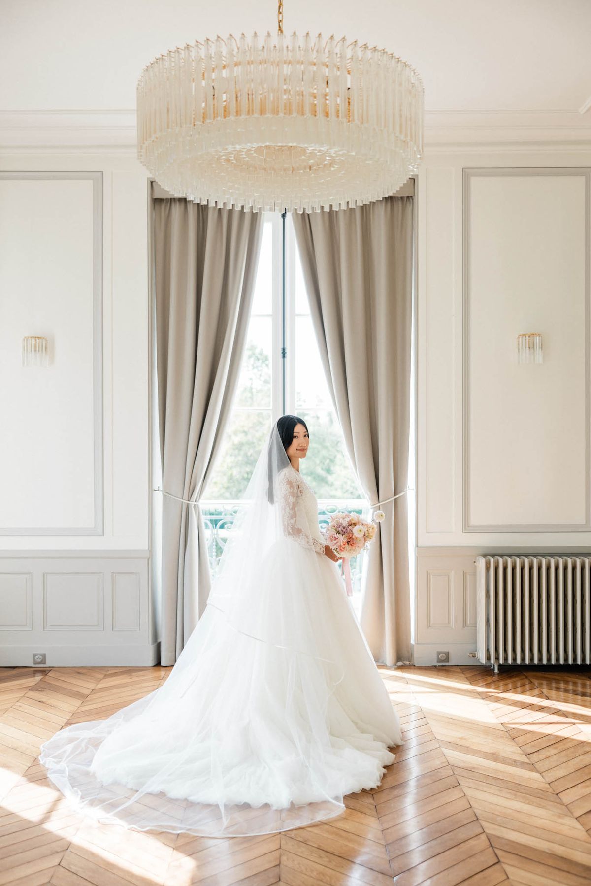Bride in lace long-sleeve ball gown with cathedral train holding a peach bouquet in a French panelled room