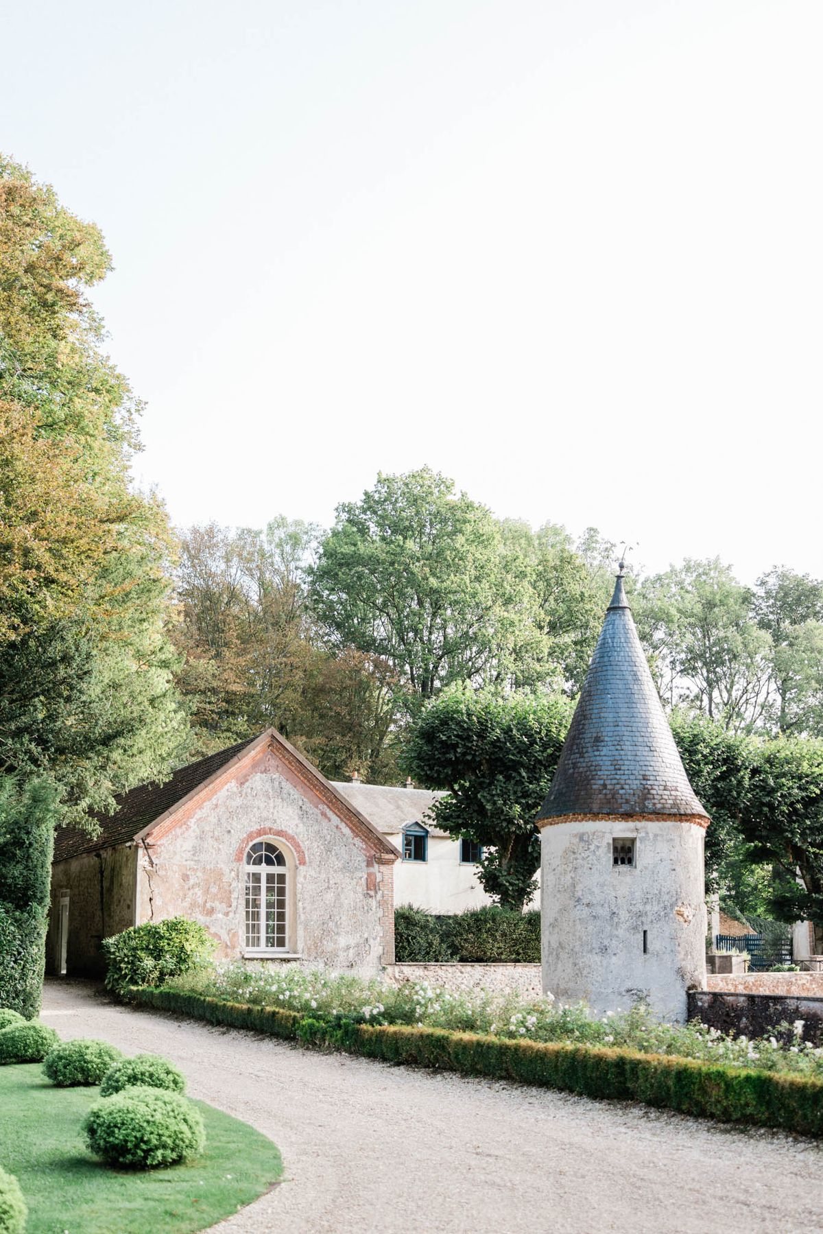 An outdoor wide shot of a French château estate featuring a weathered stone and exposed brick outbuilding with an arched white-framed window, alongside a round turret with a pointed slate roof. The grounds include a graveled driveway, neatly clipped low boxwood hedging bordering a flower bed with small white blooms, and sculpted rounded topiary shrubs on a manicured lawn in the foreground. No people are visible in the frame. The overall architectural style is classic French countryside, with the aged plaster and brick facades giving the buildings a distinctly historic character. Potential venue feature image.