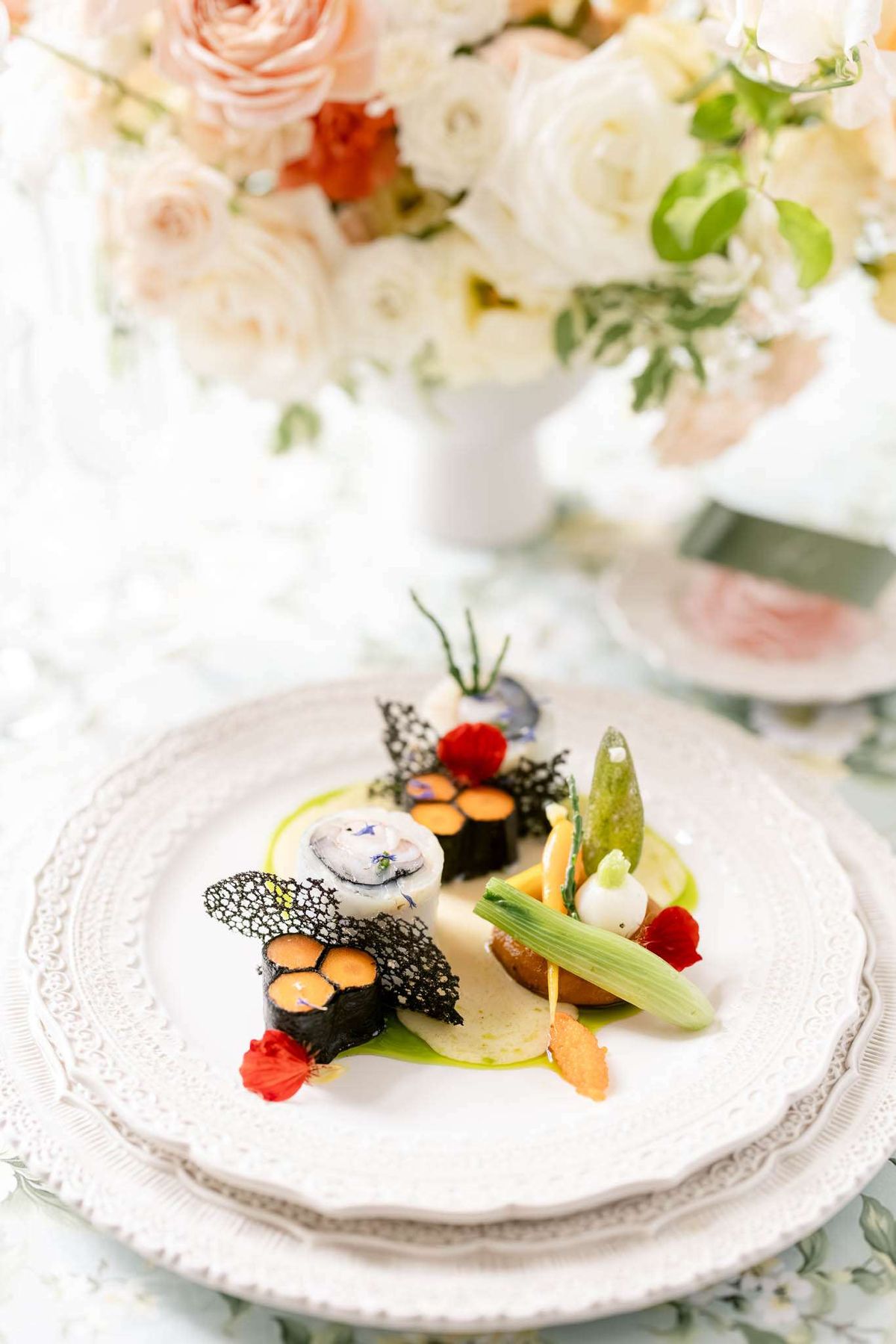 Plated starter course with seaweed rolls and edible flowers on white charger with blush rose centrepiece behind