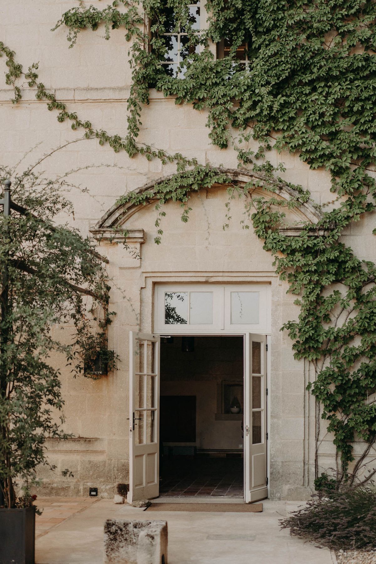 An exterior architectural detail shot of a French château or manor house, featuring a set of white French doors standing open beneath a carved stone arched surround. The cream-coloured limestone façade is heavily draped in climbing vines that trail across the arch and upper portion of the building. A transom window sits above the door panels, and a glimpse of the interior is visible through the open doors. The composition is a straight-on medium shot framing the entrance symmetrically, with no people present. Potential venue feature image.