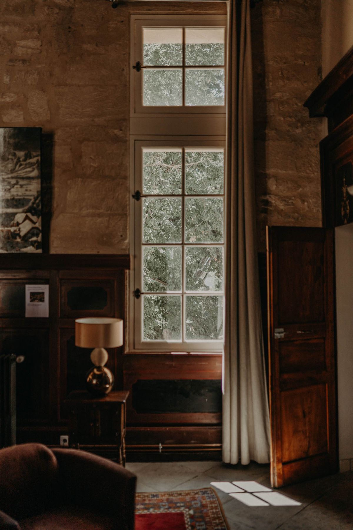 An interior room shot with no people present, showing a dimly lit space with exposed stone walls, dark wood paneling, and a tall multi-paned French window with cream linen curtains that lets in natural light. The room is furnished with antique dark wood pieces including an armoire, a side table, and a wooden chest positioned beneath the window. A gold-toned table lamp with a cream shade sits on the side table, and a patterned rug in red and muted tones is partially visible on the stone tile floor. The overall aesthetic is classic French château with warm, moody tones and period furniture. Potential venue feature image.
