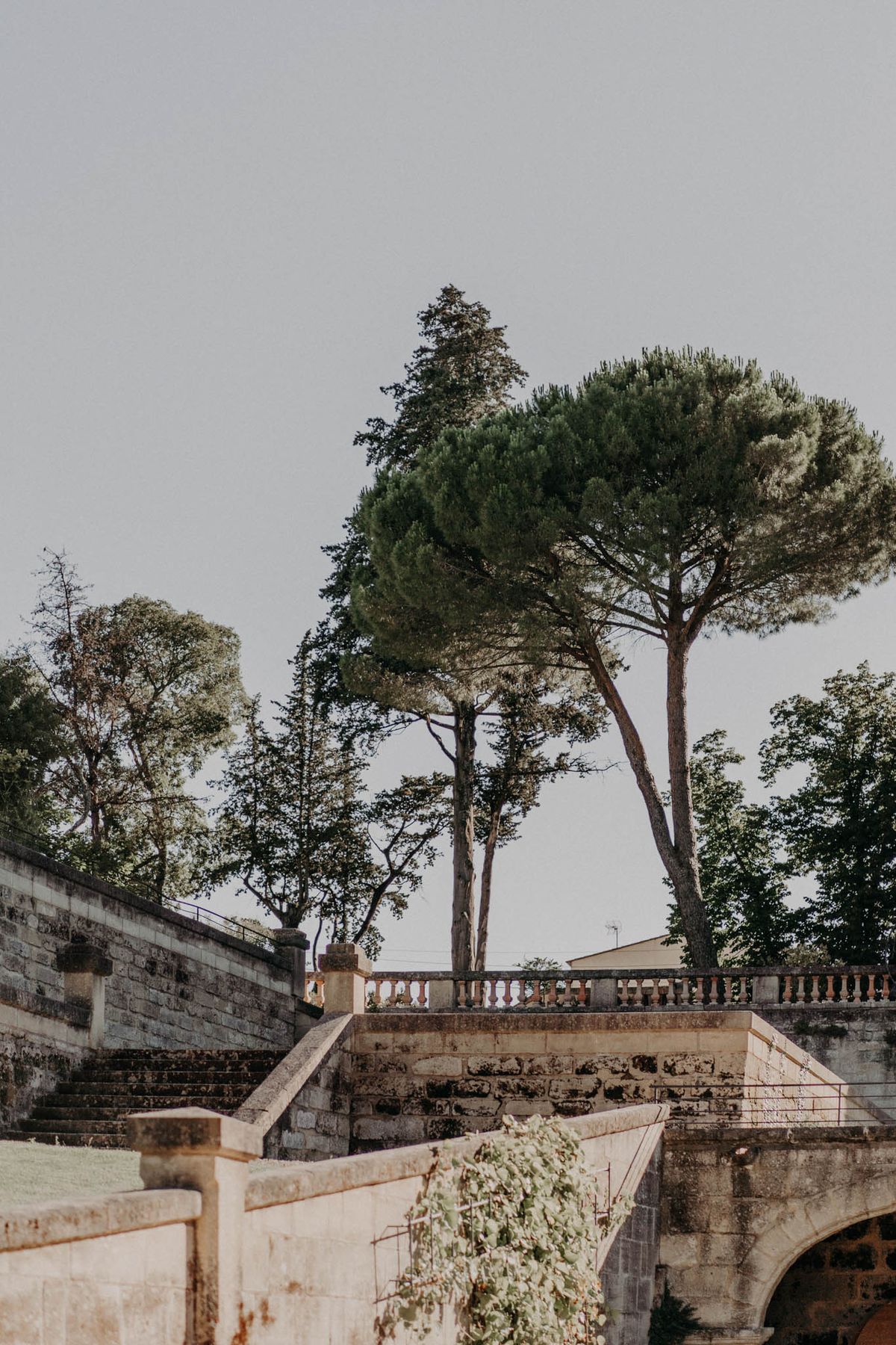 An outdoor wide shot showcasing the exterior grounds of a French château or estate, with no people visible. The image features a tiered stone terrace with a classical balustrade railing, broad stone steps ascending to an upper level, and a low arched stone structure in the foreground with trailing green foliage. The architecture is built from aged limestone blocks. The overall composition is a wide architectural shot with a muted, overcast light. Potential venue feature image.