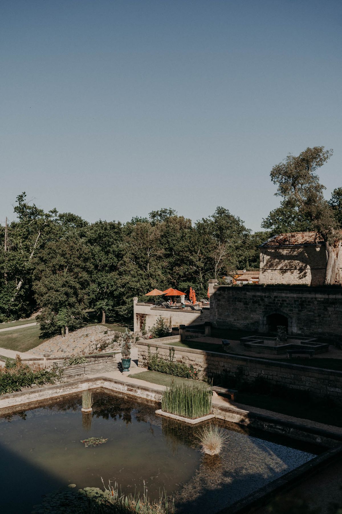 Wide outdoor shot of a French estate's grounds, showcasing formal garden terraces with a rectangular ornamental reflecting pool in the foreground, planted with aquatic grasses and lily pads in teal-glazed stone planters. The grounds are arranged across multiple stone-walled levels, with a terrace area in the middle distance featuring several burnt orange parasols and lounge seating. A historic stone outbuilding with a terracotta tile roof is visible to the right. No people are prominently visible. The overall aesthetic is classic French formal garden design with rustic stone architecture. Potential venue feature image.