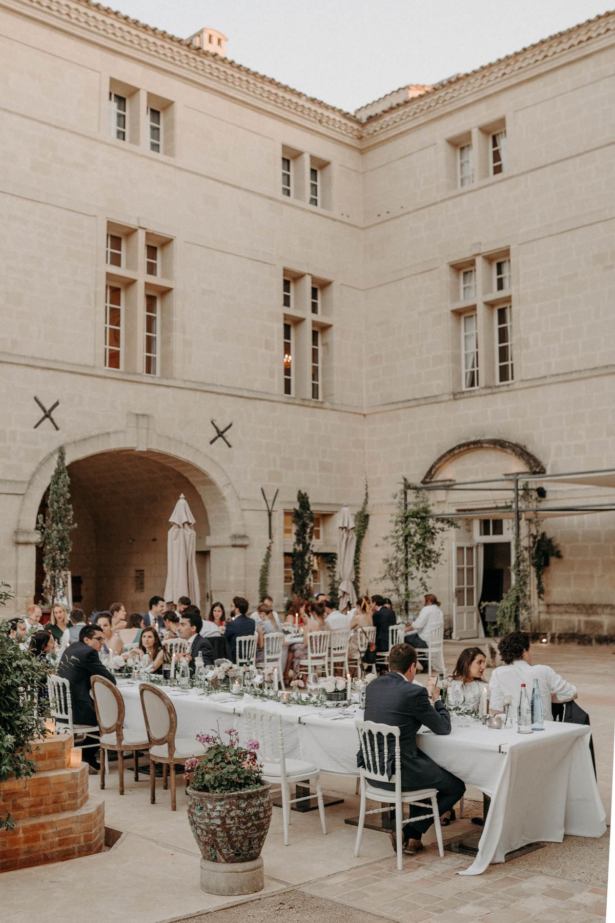 An outdoor wedding reception dinner is taking place in the interior courtyard of a large French château, with approximately 50–60 guests seated at long rectangular tables covered in white linen. The tables are set with white Chiavari and Louis XVI-style chairs, glassware, candles, and low floral centerpieces featuring light pink and white blooms with greenery. The décor palette is classic and understated, with white linens, candlelight, and trailing greenery adding softness to the stone surroundings. A large aged terracotta pot planted with pink cyclamen and trailing foliage anchors the foreground, and climbing plants frame the arched passageways of the château's façade. The image is a wide shot taken from a slightly elevated angle, capturing both the dinner scene and the full three-story limestone building behind it. Potential venue feature image.