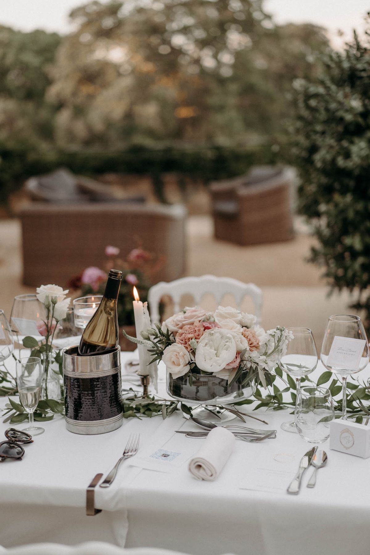 Wedding reception table with blush pink roses and white peonies centerpiece, silver candlestick, and champagne bucket at dusk