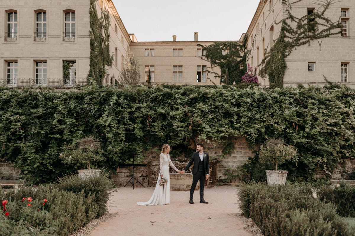 A couple portrait taken outdoors in the formal garden of a large French château, with the couple holding hands and facing each other along a gravel pathway flanked by low trimmed hedgerows and red flowering plants. The bride wears a long-sleeved ivory lace gown with a flowing skirt and train, and carries a loose bouquet with warm-toned blooms. The groom wears a dark navy three-piece suit with a white dress shirt and a small floral buttonhole. Behind them, a tall dense hedge of climbing greenery lines a stone wall, with the multi-story limestone château building visible above, partially covered in ivy with hints of purple flowering vines on the right wing. The image is a wide shot with the couple centered at mid-distance, giving prominence to both the architecture and the formal garden layout. Potential venue feature image.