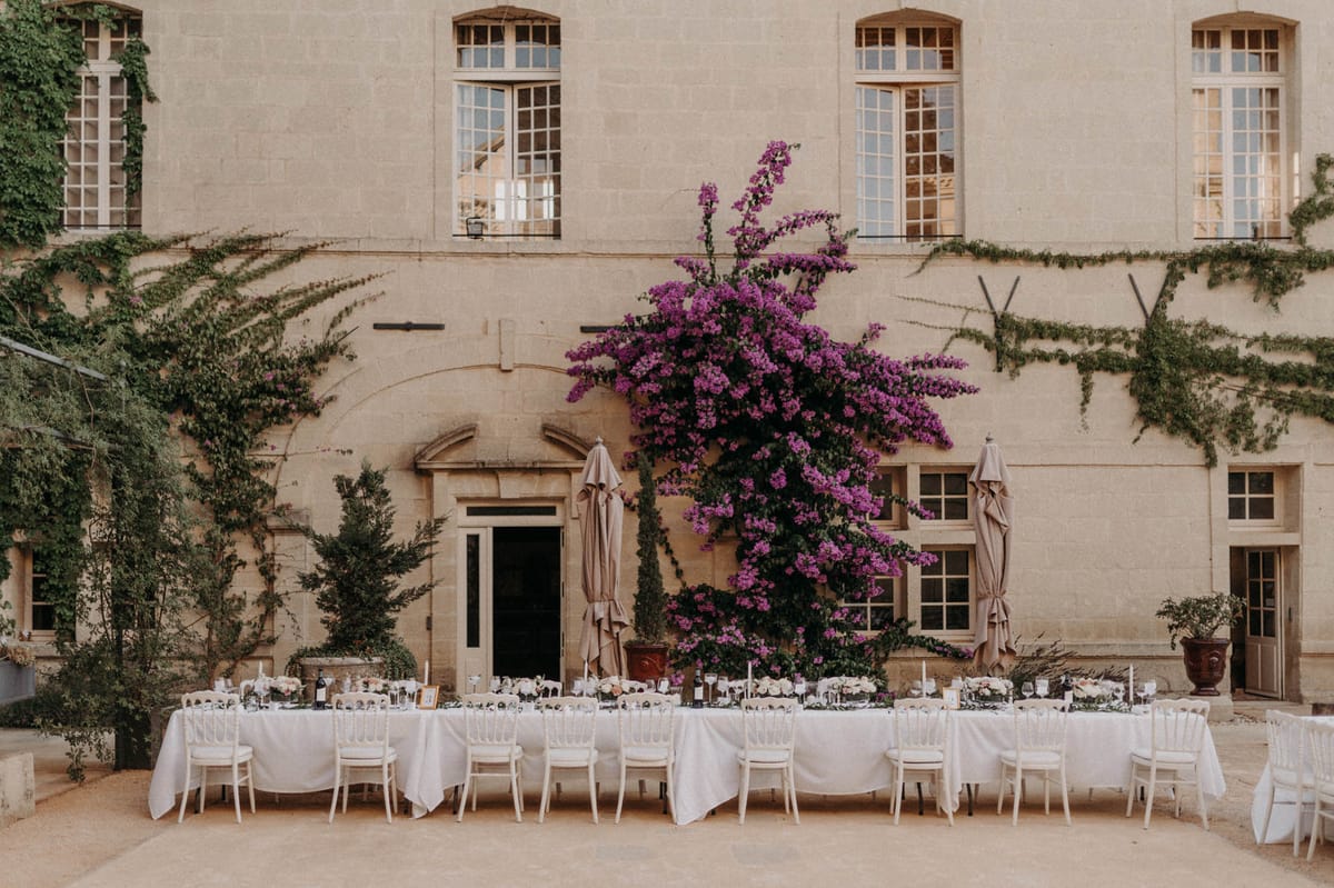 An outdoor wedding reception setup in the courtyard of a French château, featuring two long banquet tables dressed in white linen tablecloths with white Napoleon chairs arranged along both sides. The tables are set with glassware, candlesticks, and low floral centerpieces in soft ivory and blush tones with greenery. The setting is framed by the château's pale stone façade with tall multi-pane windows, and a large bougainvillea in vivid magenta-purple dominates the center of the composition, climbing the wall above a classical arched doorway. Two taupe closed parasols flank the entrance, and potted topiaries and urns add symmetry to the scene. The overall decor palette is white, ivory, and soft blush with the bougainvillea providing a bold color contrast. Wide establishing shot with no people present. Potential venue feature image.