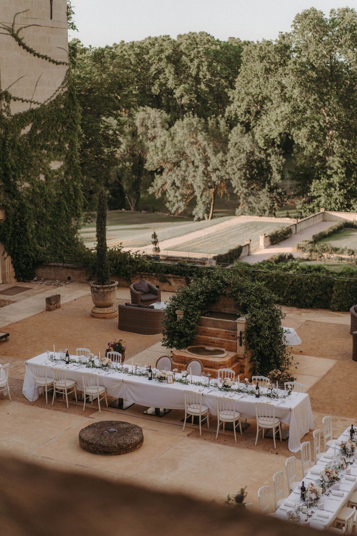 An elevated wide shot of an outdoor wedding reception setup on a stone-paved terrace, likely at a French château. Multiple long banquet tables are dressed in white linen tablecloths and lined with white Chiavari-style chairs, with garlands of greenery and small floral arrangements in peach, blush, and purple tones running along the centers. A large stone fountain overgrown with a dense leafy climber sits at the heart of the layout, acting as a natural focal point between the tables. The terrace overlooks a formal French garden with clipped hedges and a rectangular reflecting pool or parterre in the background. The decor palette is classic and natural, combining white linens, lush greenery, and soft floral tones in a style that reads as refined outdoor dining. Potential venue feature image.
