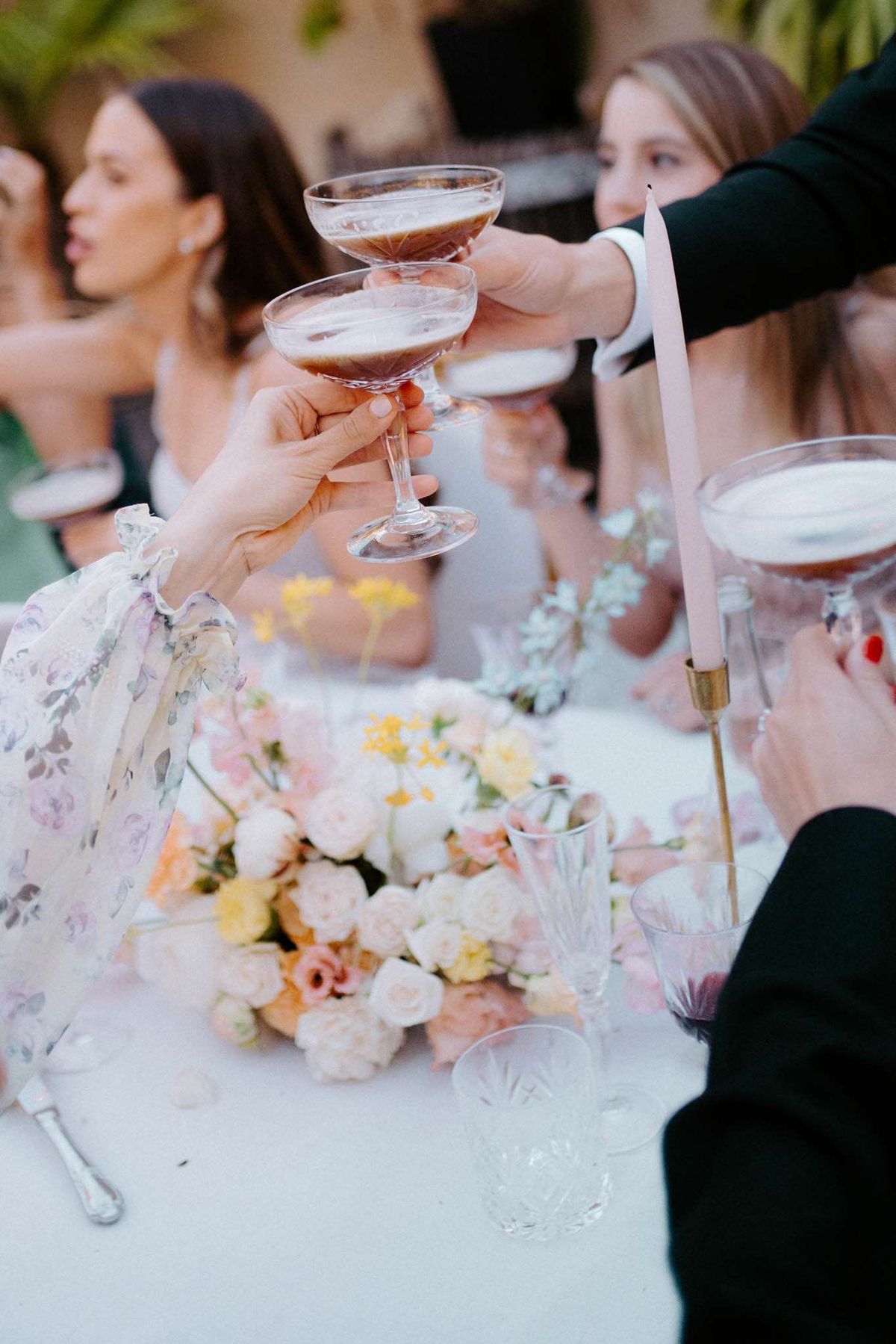 Guests are raising coupe glasses filled with what appears to be espresso martinis in a toast during a wedding reception. The shot is taken at an outdoor table set with a white linen, a low centerpiece of cream, blush, peach, and yellow roses, and a slender blush-pink taper candle in a gold candleholder. Cut crystal glassware and a wine glass with red wine are also visible on the table. In the foreground, one guest wears a floral-print organza dress with ruffled sleeves in soft pink and lavender tones, while other guests in the background wear light grey and dark outfits. The composition is a close-up, shallow-depth-of-field shot focused on the clinking coupes, with guests softly blurred behind. The overall decor palette is soft and garden-inspired, mixing pastel florals with classic crystal and brass accents.