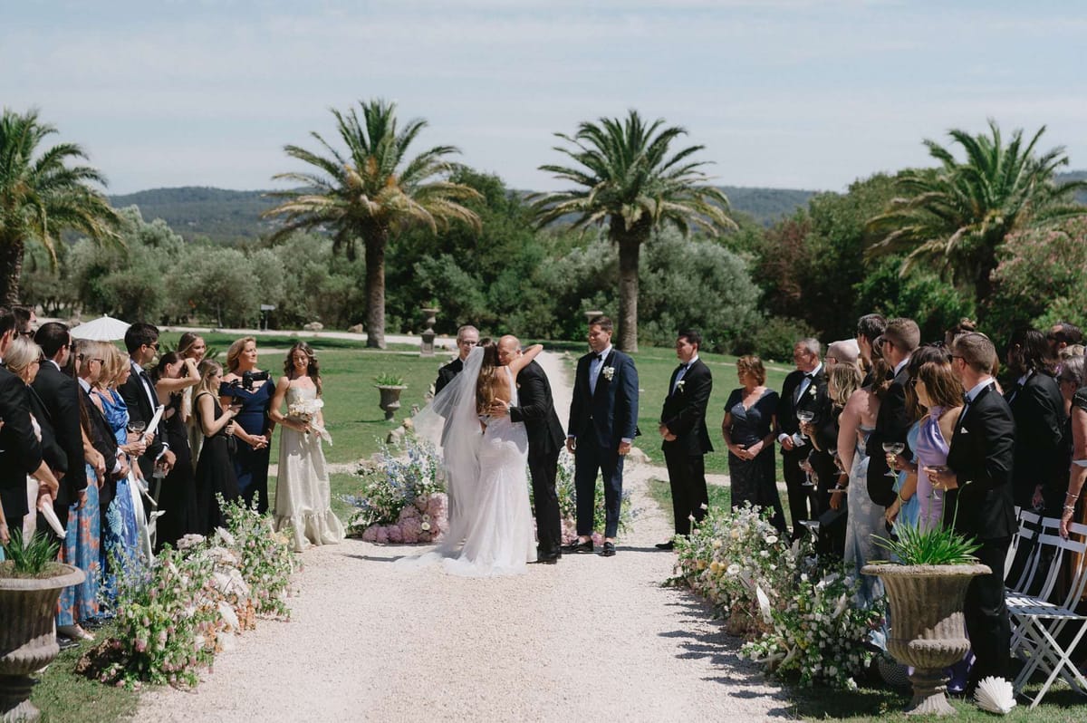 An outdoor wedding ceremony kiss moment captured on a gravel allée lined with guests on both sides, set within a French estate grounds with tall palm trees and rolling hills in the background. The bride wears a fitted white lace gown with a long veil and carries a bouquet, while the groom is in a dark navy suit; a best man in a navy suit stands to the right of the couple. The aisle is decorated with low garden-style floral arrangements featuring soft pink, blush, dusty blue, and ivory blooms — including what appear to be delphiniums, roses, and ranunculus — spilling naturally along the gravel, with large stone urns anchoring the arrangements. Approximately 40–50 guests line the aisle, dressed in formal attire including black tuxedos, navy and black evening gowns, and a bridesmaid in an ivory tiered dress; white folding chairs are visible at the edges. The wide-shot composition captures the full ceremony scene from behind the couple, emphasizing the length of the floral-lined aisle and the open landscape beyond.
