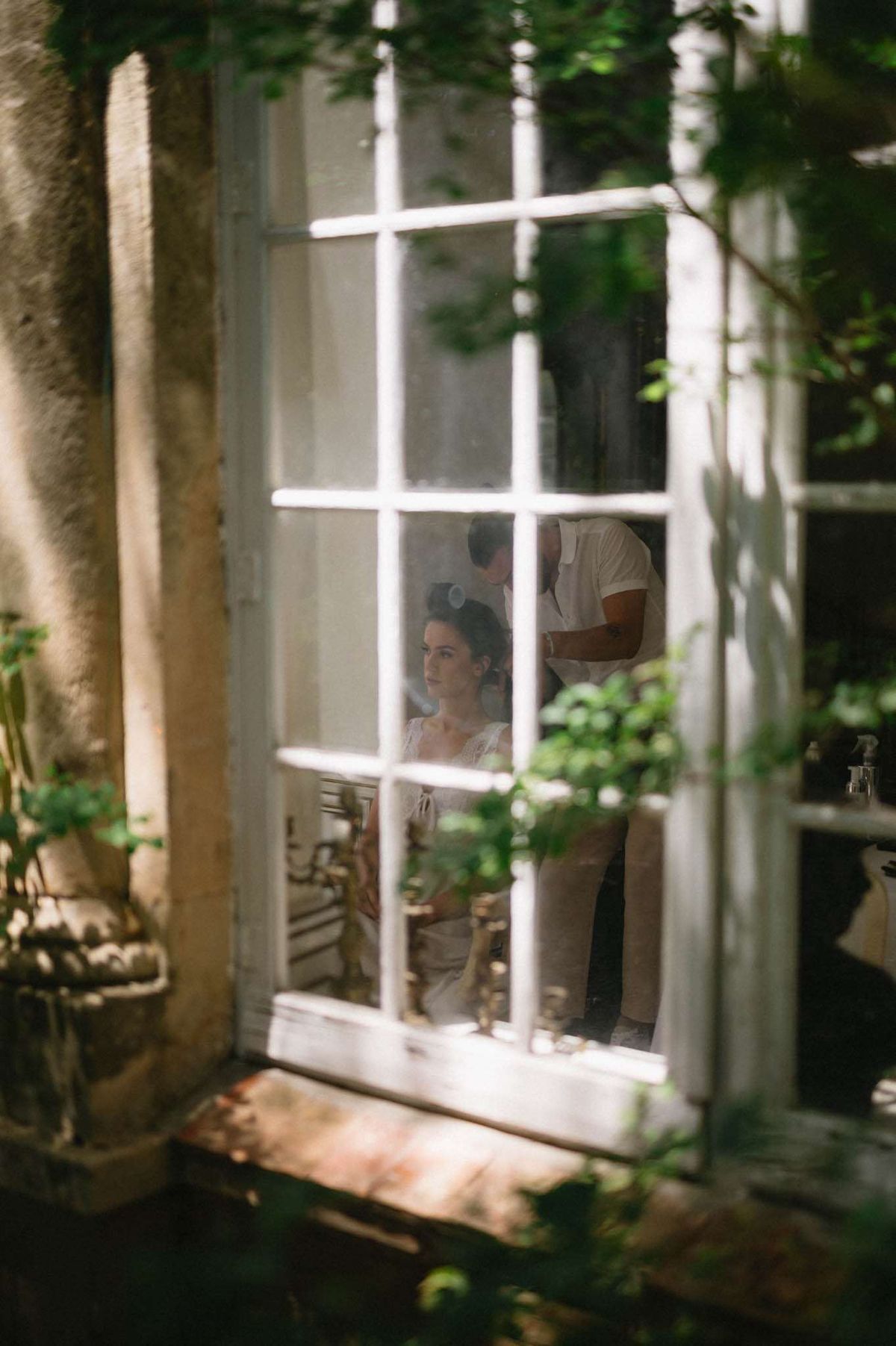 Getting ready scene shot through French window panes showing bride with hair stylist
