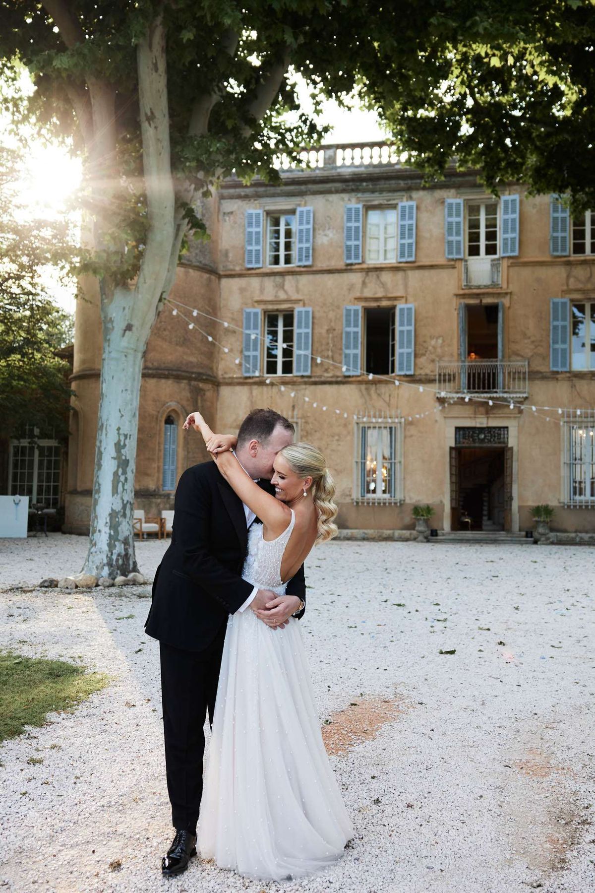 A couple portrait shot outdoors on the gravel forecourt of a large French château at golden hour, with warm sunlight flaring through the tree canopy to the left. The groom, wearing a classic black tuxedo with a bow tie and black dress shoes, leans in to kiss the bride's cheek while holding her close with one arm around her waist; she has her arm raised around his neck and is smiling. The bride wears a white spaghetti-strap gown with an open back and a softly embellished skirt, with her blonde hair styled in loose waves over one shoulder. The château facade behind them features warm ochre-toned render, pale blue shutters across multiple floors, a wrought-iron balcony, and strings of bistro lights strung across the courtyard. The composition is a full-length portrait with the venue prominently featured in the background. Potential venue feature image.