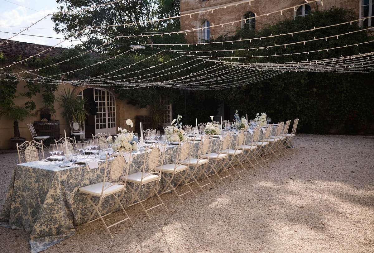 Toile de Jouy table with blue hydrangeas white roses silver candlesticks wrought-iron rattan chairs under fairy light canopy