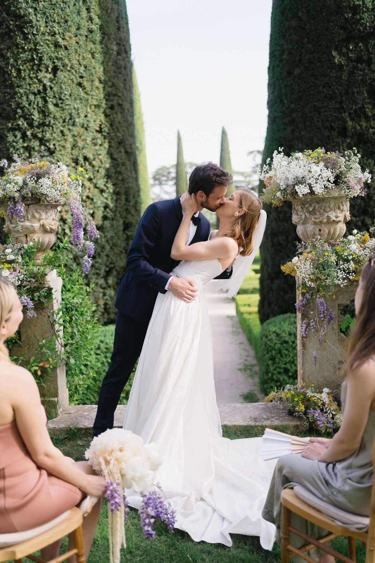 Bride and groom share first kiss between stone urns with wildflower arrangements in formal hedged garden