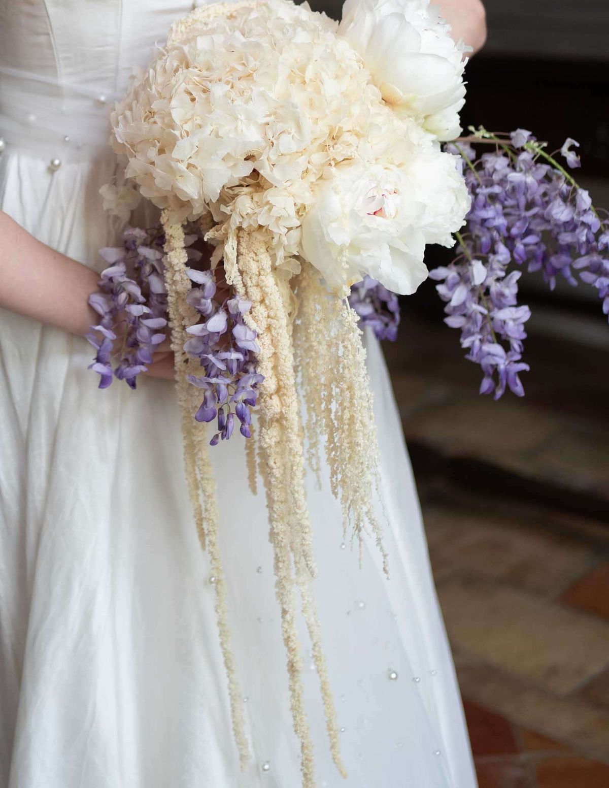 Cascading bouquet of cream peonies, trailing amaranthus, and purple wisteria held by bride in pearl-embellished gown