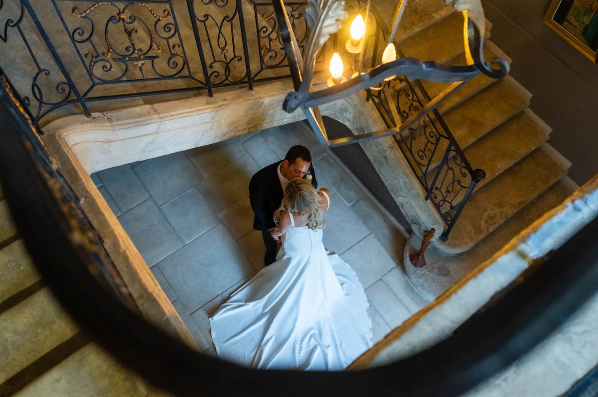A couple shares a kiss at the base of a grand interior stone staircase, photographed from directly above in a wide overhead shot. The bride wears a white satin gown with a long train and button-detail along the back, with her blonde hair styled up, while the groom wears a dark navy suit with a white boutonnière. The staircase features ornate wrought-iron balustrades with scrollwork detailing, and the stone steps and landing have a warm honey tone. A chandelier with candle-style bulbs is partially visible in the foreground, adding warm amber light to the scene. The interior setting suggests a classic French château, and the composition frames the couple within the geometry of the stairwell. Potential venue feature image.